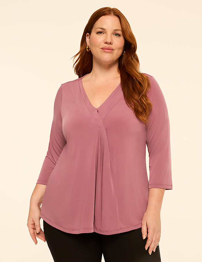 A woman with auburn hair and a mauve v-neck top stands against a plain cream background.