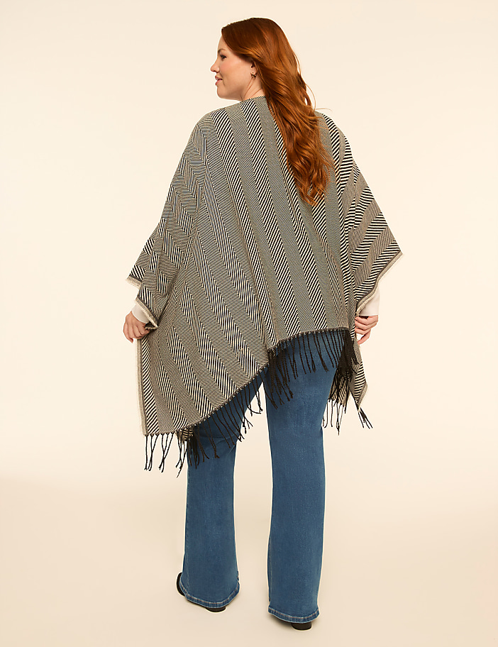 A woman with red hair stands facing away from the camera, wearing a patterned poncho and flared jeans against a plain, light-colored background.