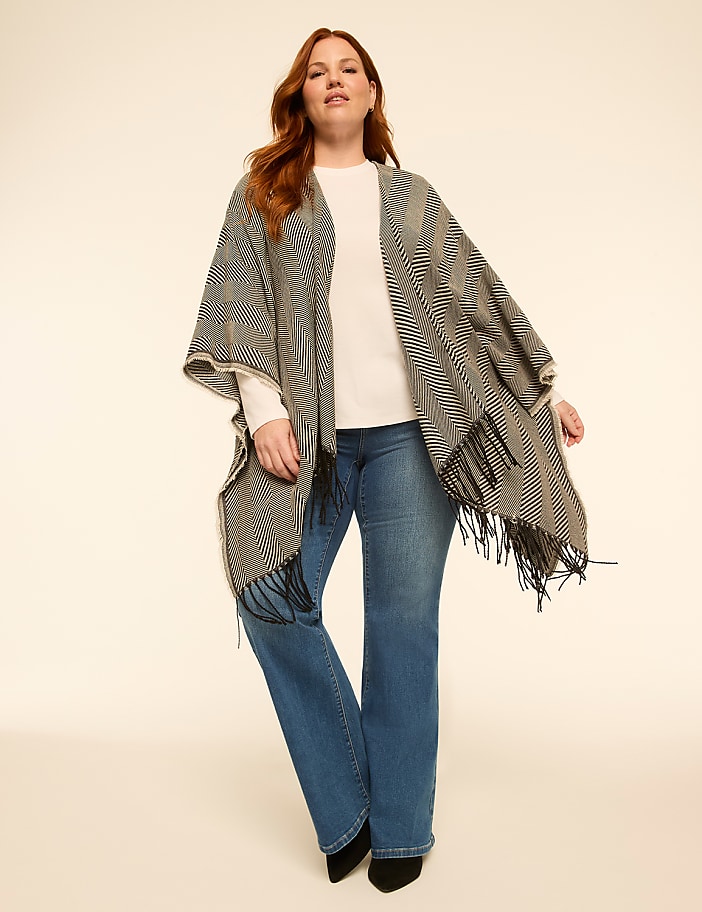 A woman in a patterned poncho and jeans stands against a plain, light-colored background.