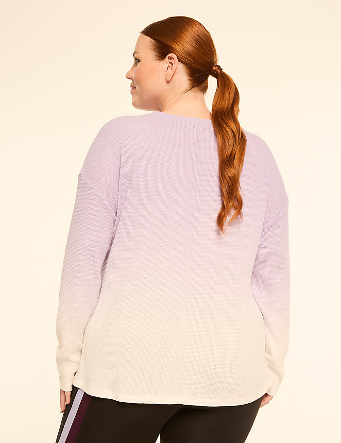 A woman with red hair pulled into a ponytail wears a light purple and cream ombre waffle-knit sweater and black pants with purple and white stripes, against a plain cream background.