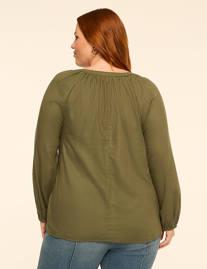 A woman with red hair wearing a green blouse and blue jeans stands against a plain, light-colored background.