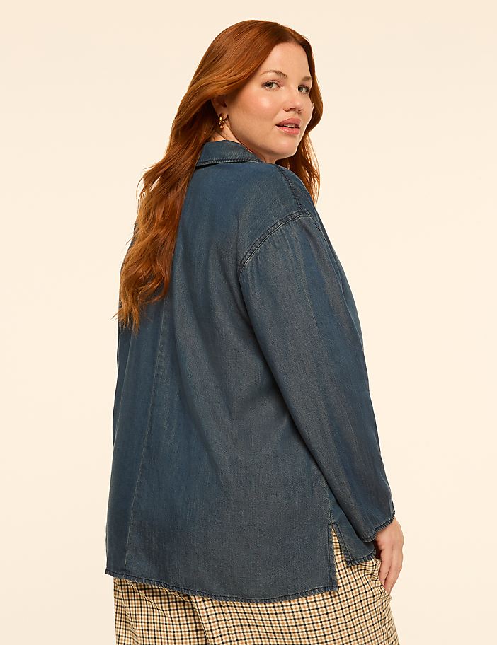 A woman with long red hair wearing a denim shirt and plaid pants stands against a plain, light-colored background.