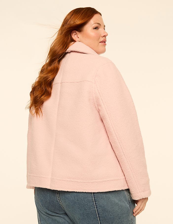 A woman with long, wavy red hair wearing a pink jacket and blue jeans stands against a plain, light-colored background.