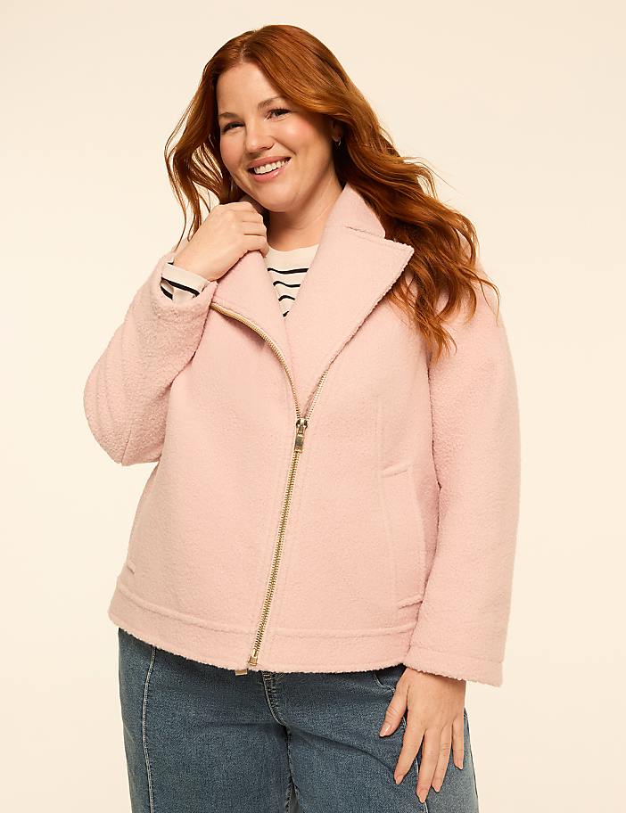 A smiling woman with red hair in a pink jacket and jeans stands against a plain cream background.