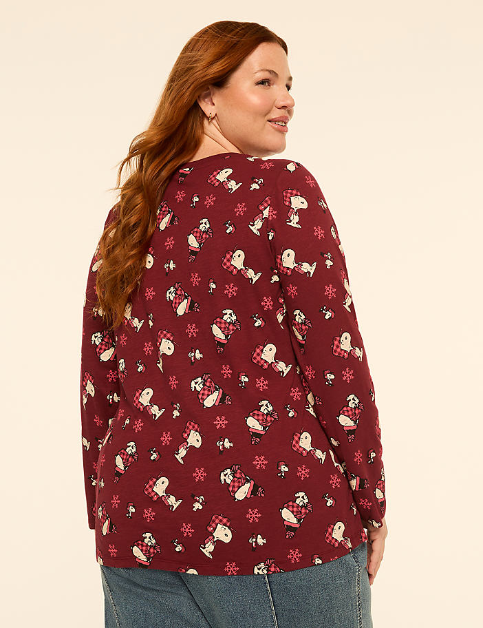 A woman with red hair wearing a maroon long-sleeved shirt with a Snoopy pattern stands against a plain, light-colored background.