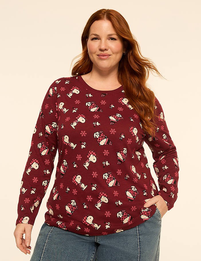 A woman with red hair and a patterned long-sleeved shirt stands against a plain, light-colored background.