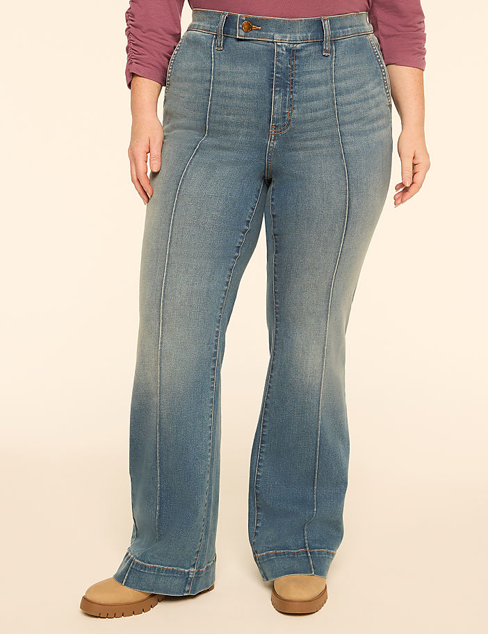 A person wearing flared jeans and tan shoes stands against a plain, light-colored background.