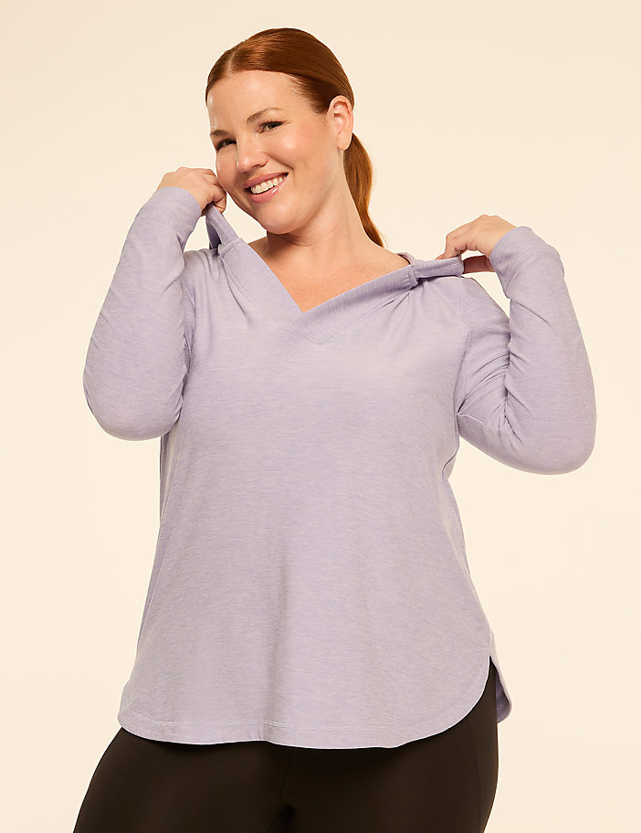 A smiling woman in a light purple hoodie and black leggings stands against a plain, light beige background.