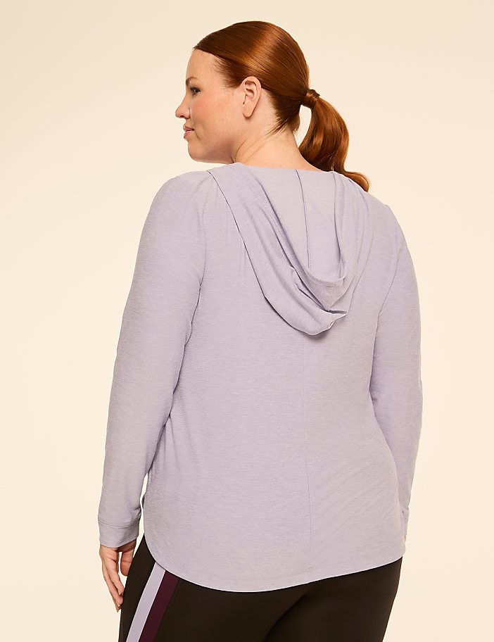 A woman with auburn hair pulled into a ponytail wears a light purple hooded shirt and black leggings, standing against a plain, light beige background.