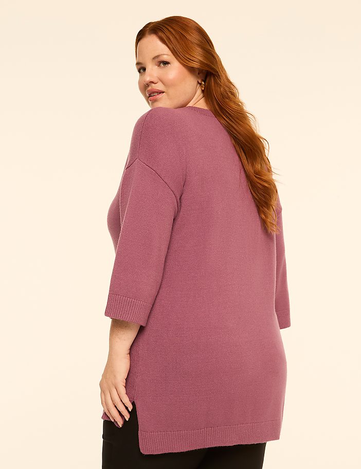 A woman with red hair wearing a mauve sweater stands against a plain, light-colored background.