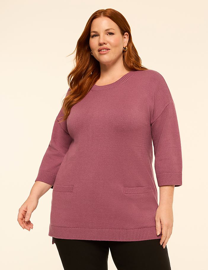 A woman with red hair stands in the foreground wearing a mauve sweater with pockets, against a plain, light-colored background.