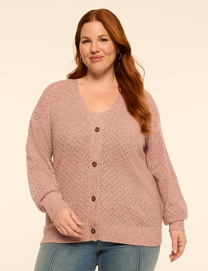A woman with red hair smiles in a pink cardigan and jeans against a plain, light-colored background.