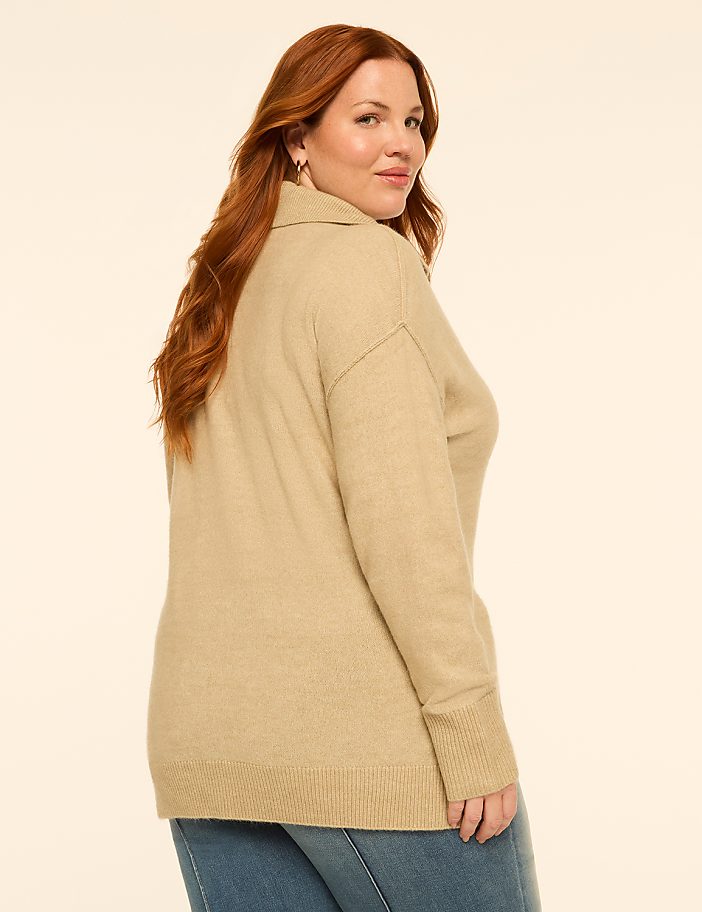 A woman with red hair wearing a tan turtleneck sweater and blue jeans poses against a plain, light-colored background.