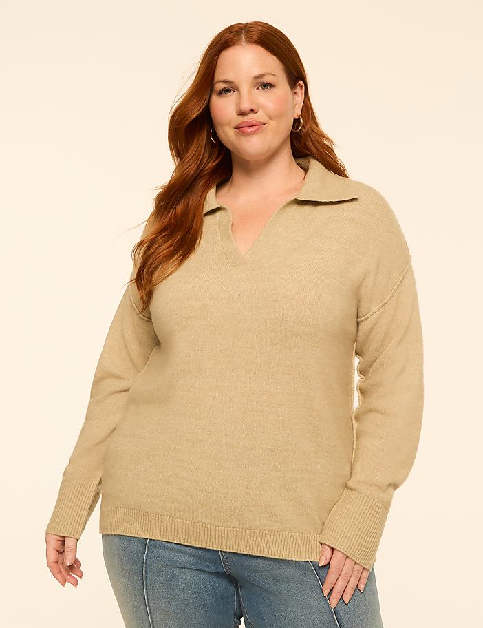 A woman with red hair and fair skin, wearing a tan collared sweater and blue jeans, stands against a plain, light-colored background.