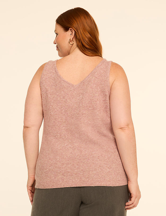 A woman with auburn hair, wearing a pink V-neck tank top and dark green pants, stands against a plain, light-colored background.