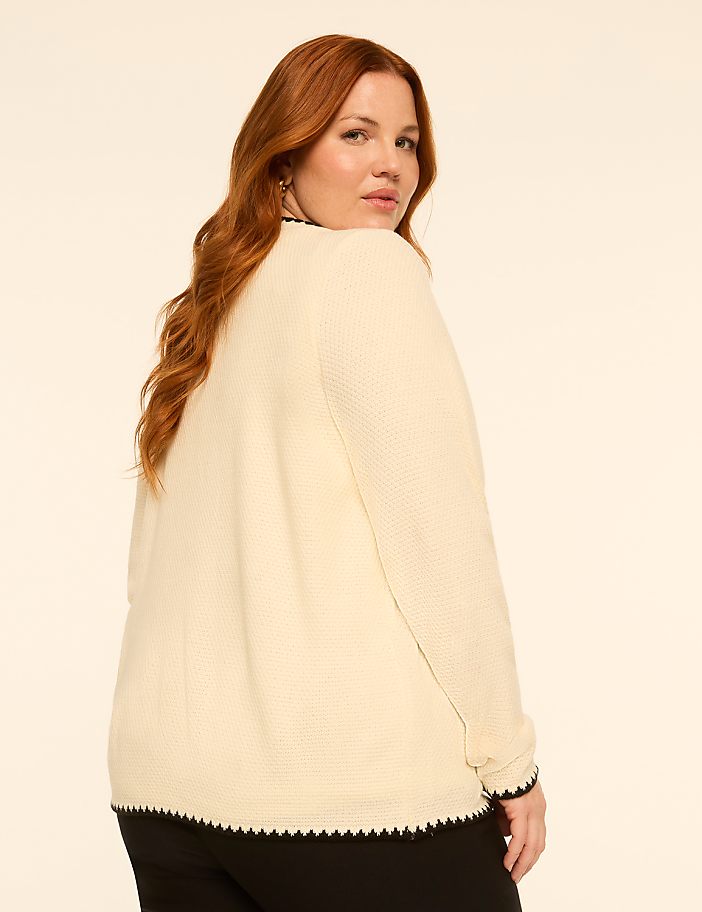 A woman with red hair, wearing a cream-colored cardigan with black trim, poses against a plain, light-colored background.