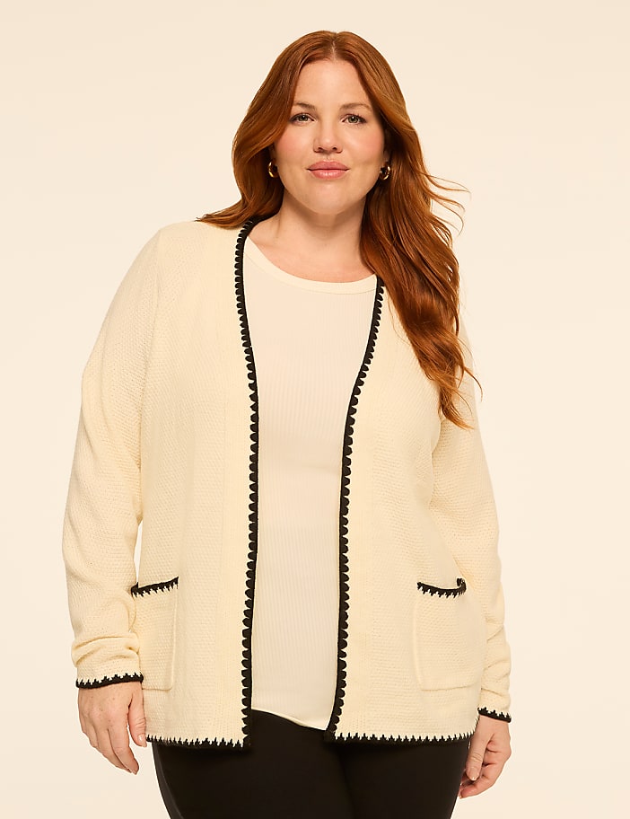 A woman with auburn hair stands in the foreground wearing a cream-colored cardigan with black trim, against a plain, light-colored background.