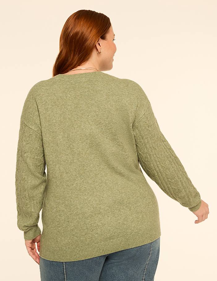 A woman with red hair wearing a green cable-knit sweater and blue jeans stands against a plain, light-colored background.