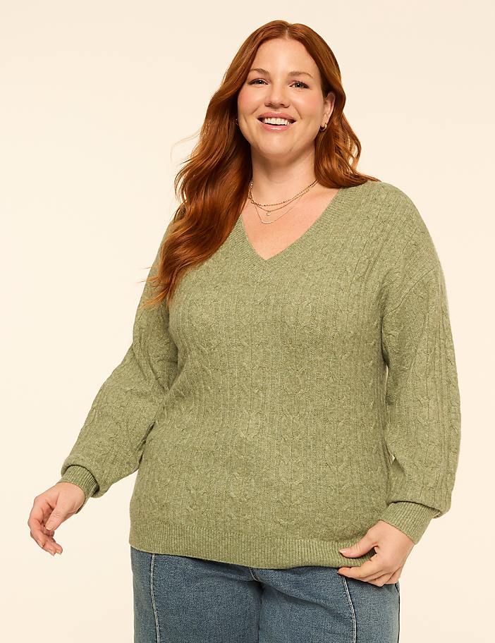 A smiling woman with red hair wears a green cable-knit sweater and blue jeans against a plain, light-colored background.
