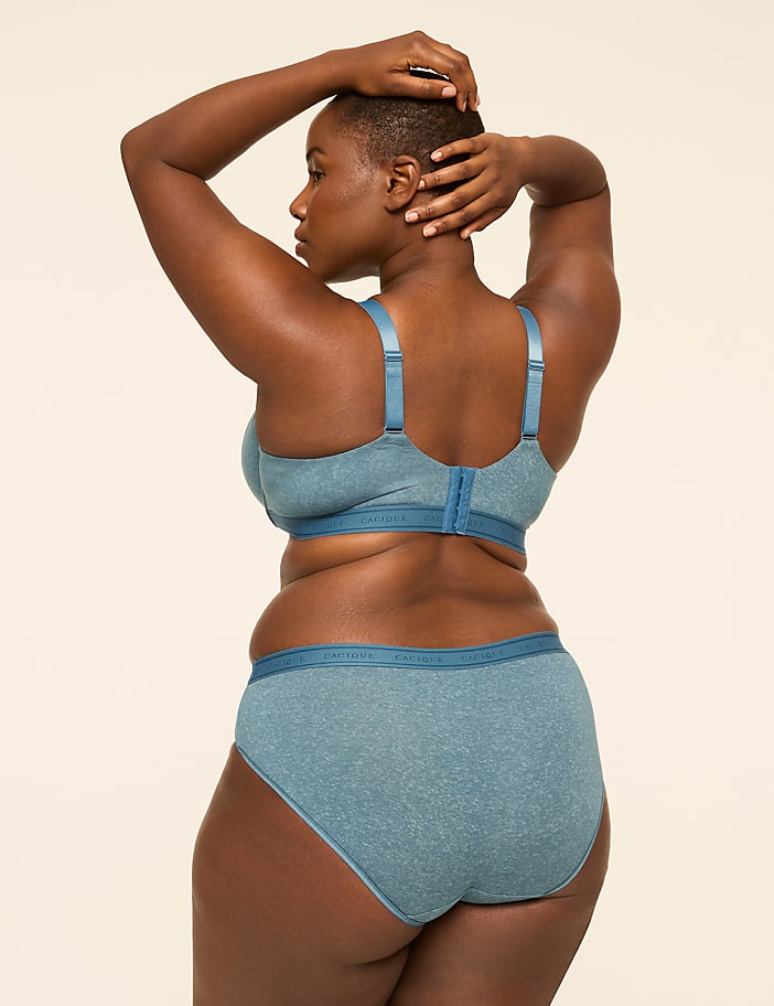 A woman in blue lingerie stands with her hands behind her head against a plain, light-colored background.