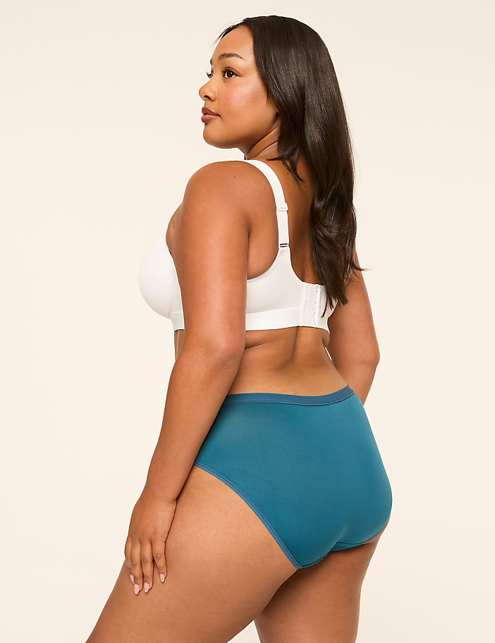 A woman in a white bra and teal underwear stands against a plain, light background.