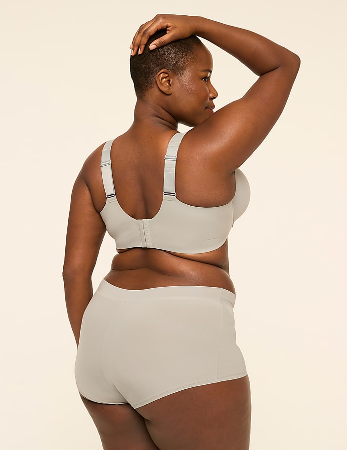 A woman in a beige bra and underwear stands with her hand on her head against a plain, light-colored background.