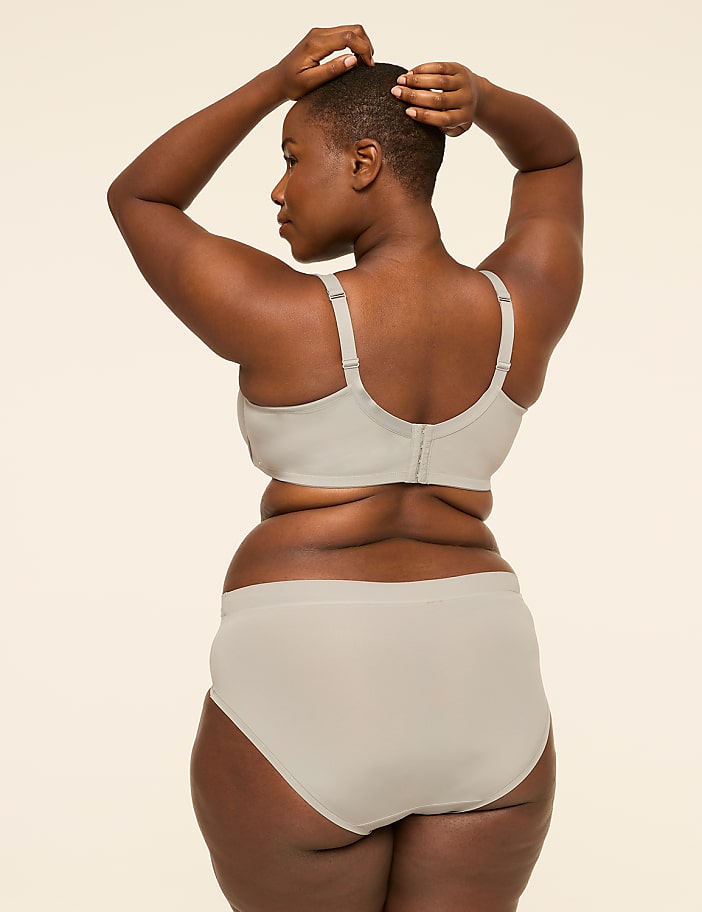 A woman in a light gray bra and underwear stands with her back to the viewer, hands on her head, against a plain, light background.