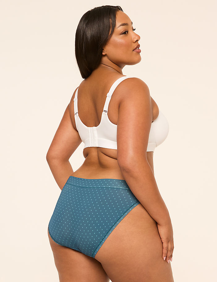 A woman in a white bra and teal polka-dot underwear stands against a plain, light brown background.