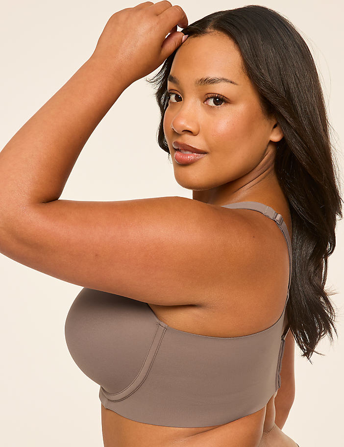 A woman in a taupe bra poses with her hand on her head against a plain, light background.