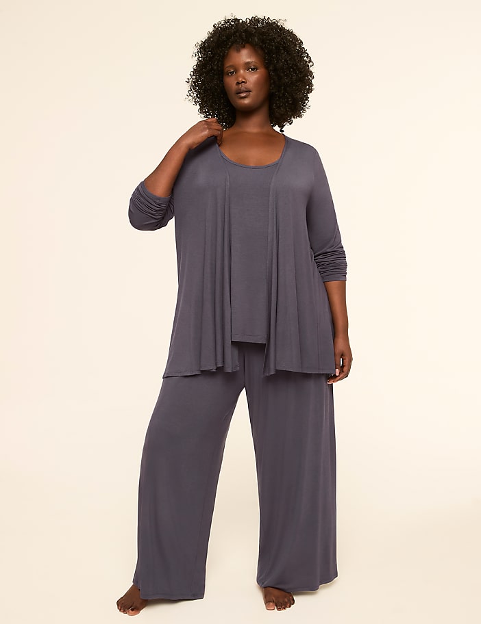A woman with dark, curly hair stands in a grey loungewear set against a plain, light background.