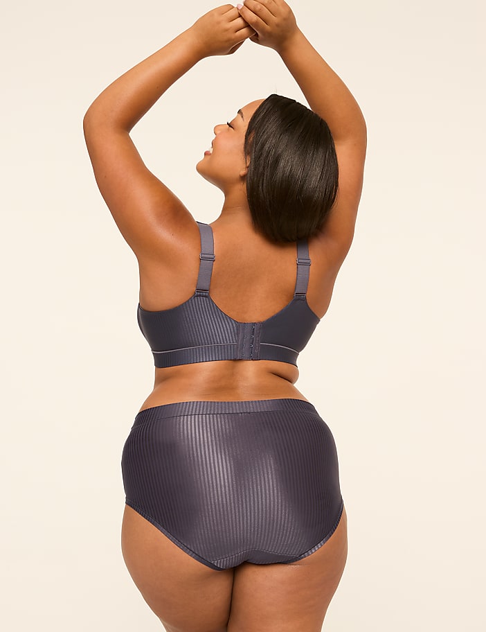 A woman in a grey ribbed bra and high-waisted briefs poses with her arms raised against a plain, light-colored background.