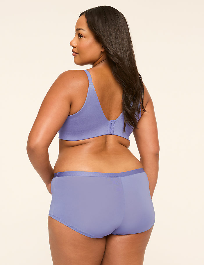 A woman in a lavender bra and boy shorts stands with her back to the viewer against a plain, light-colored background.