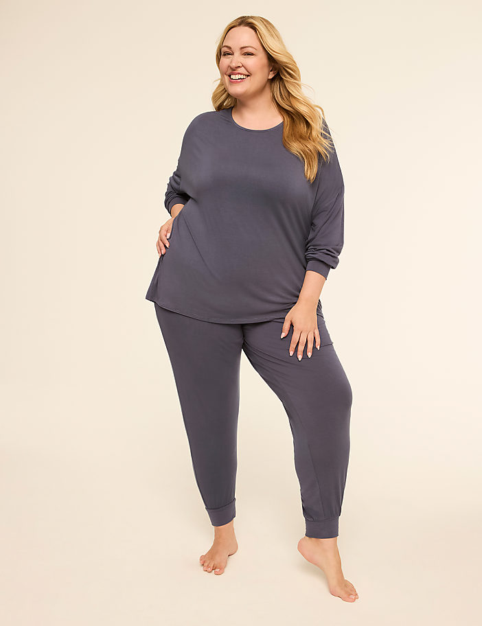 A smiling woman in a grey pajama set stands on a plain, light-colored floor against a soft, neutral background.