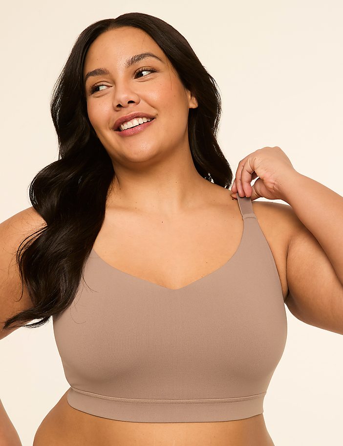 A smiling woman with dark hair wears a beige bra in the foreground against a plain, light brown background.