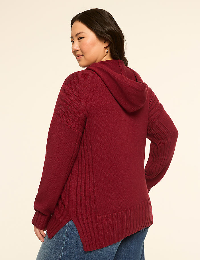A woman in a maroon hooded sweater and blue jeans stands against a plain, light-colored background.