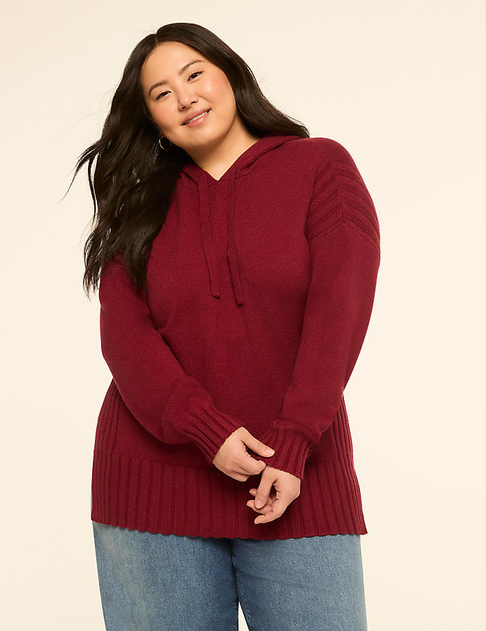 A smiling woman in a maroon hooded sweater and blue jeans stands against a plain cream background.