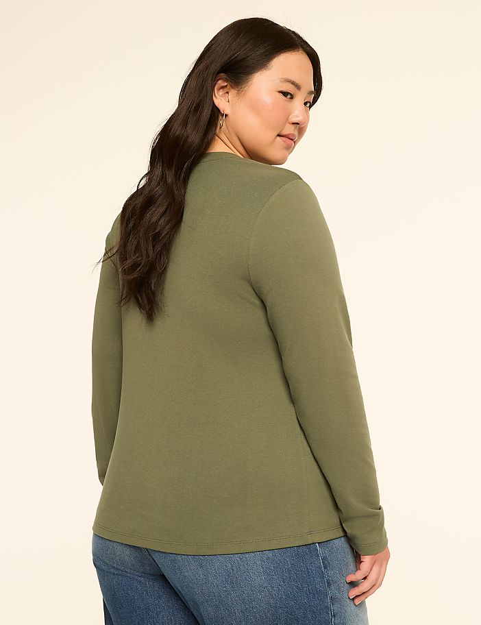 A woman in a green long-sleeved shirt and blue jeans stands against a plain, light-colored background.