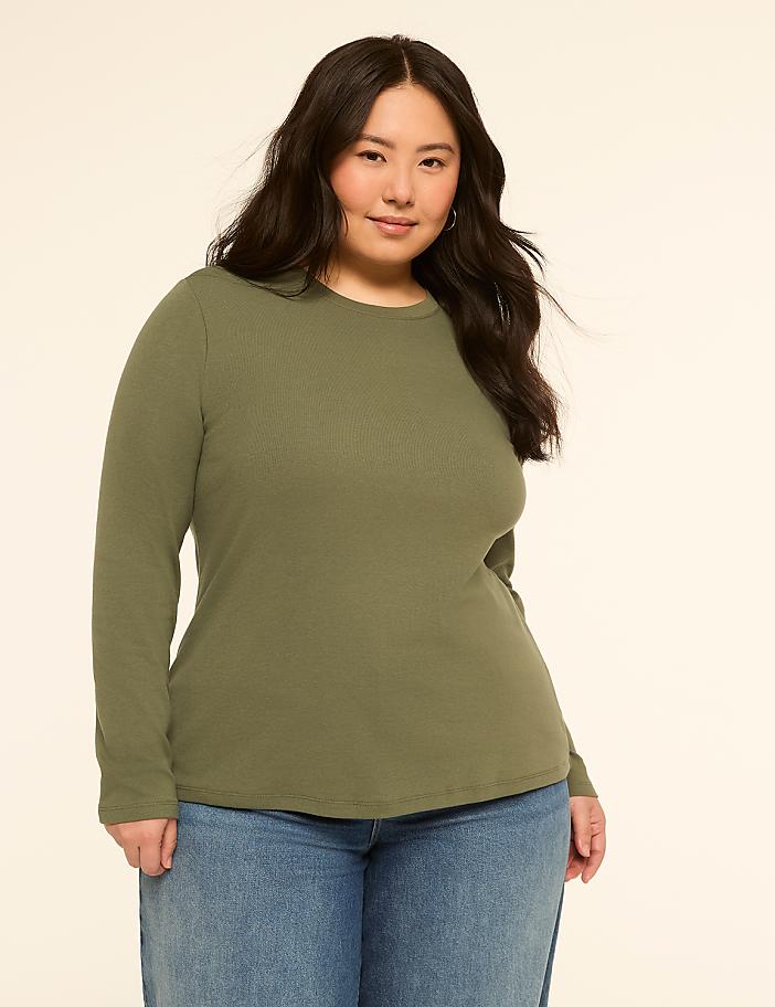 A woman with dark hair and a warm smile poses in a green long-sleeved shirt and blue jeans against a soft, neutral background.