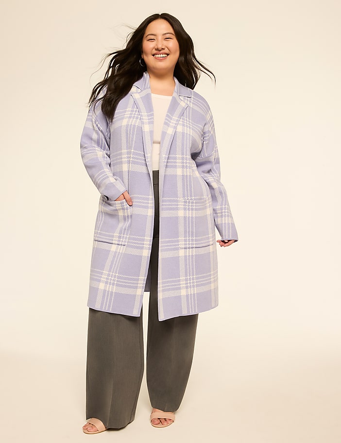A smiling woman in a light purple plaid coat and gray pants stands against a plain, light-colored background.