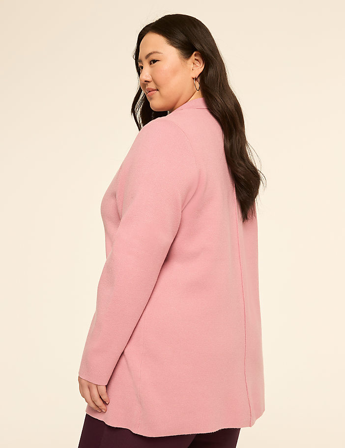 A woman in a pink cardigan and dark pants stands against a plain, light-colored background.