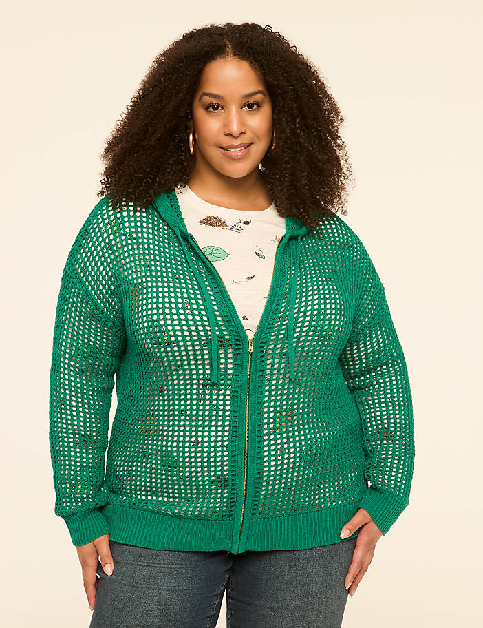 A smiling woman with curly hair wears a green hooded cardigan over a patterned shirt and jeans, standing against a plain, light-colored background.