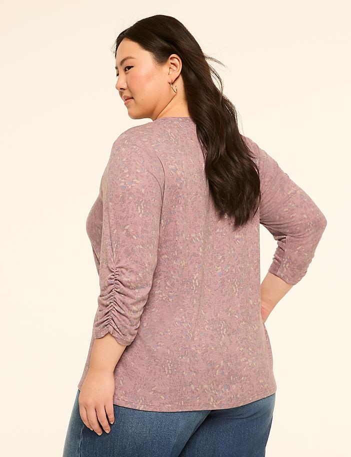 A woman in a textured pink top and blue jeans stands against a plain, light-colored background.