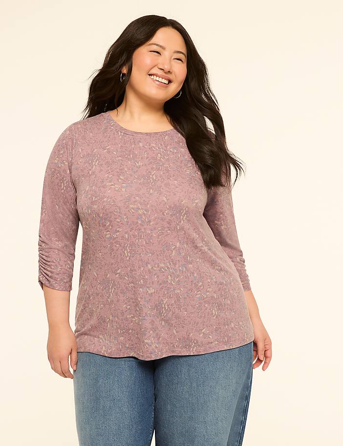 A smiling woman in a patterned pink shirt and blue jeans stands against a plain cream background.