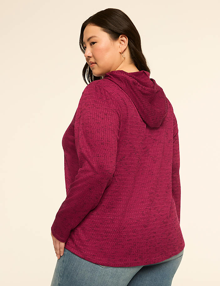 A woman in a magenta hooded sweater and jeans stands against a plain, light-colored background.