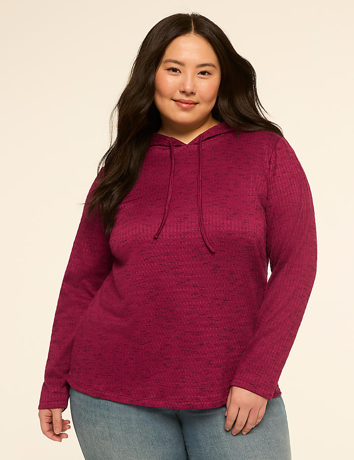 A woman in a magenta hooded sweatshirt and blue jeans stands against a plain, light-colored background.