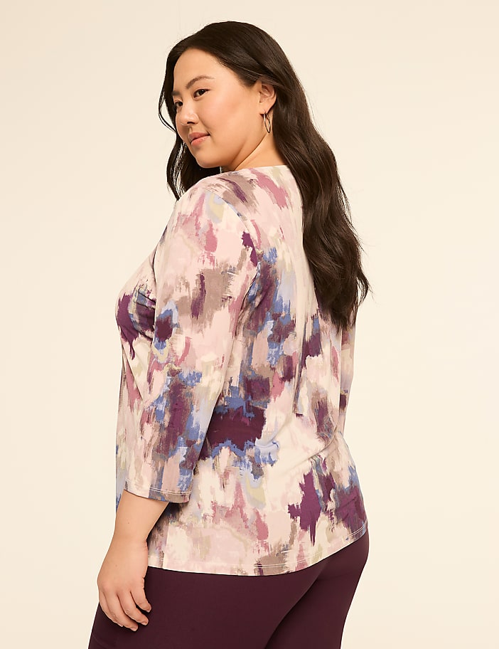A woman in a colorful abstract print top and dark pants poses against a plain, light-colored background.