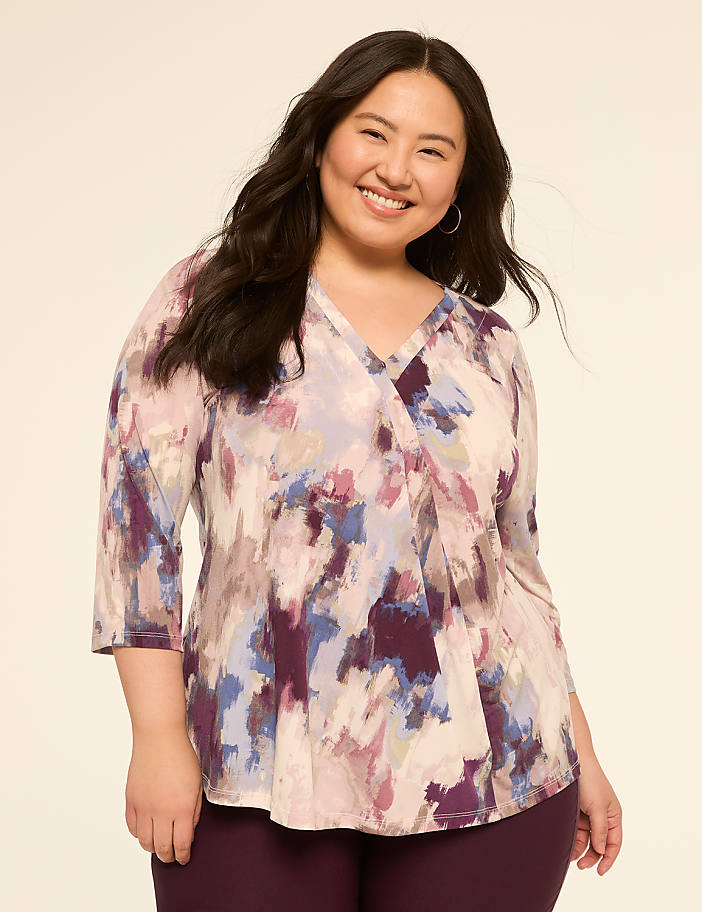A smiling woman with dark hair wears a colorful, abstract-print blouse and dark pants against a plain, light-colored background.