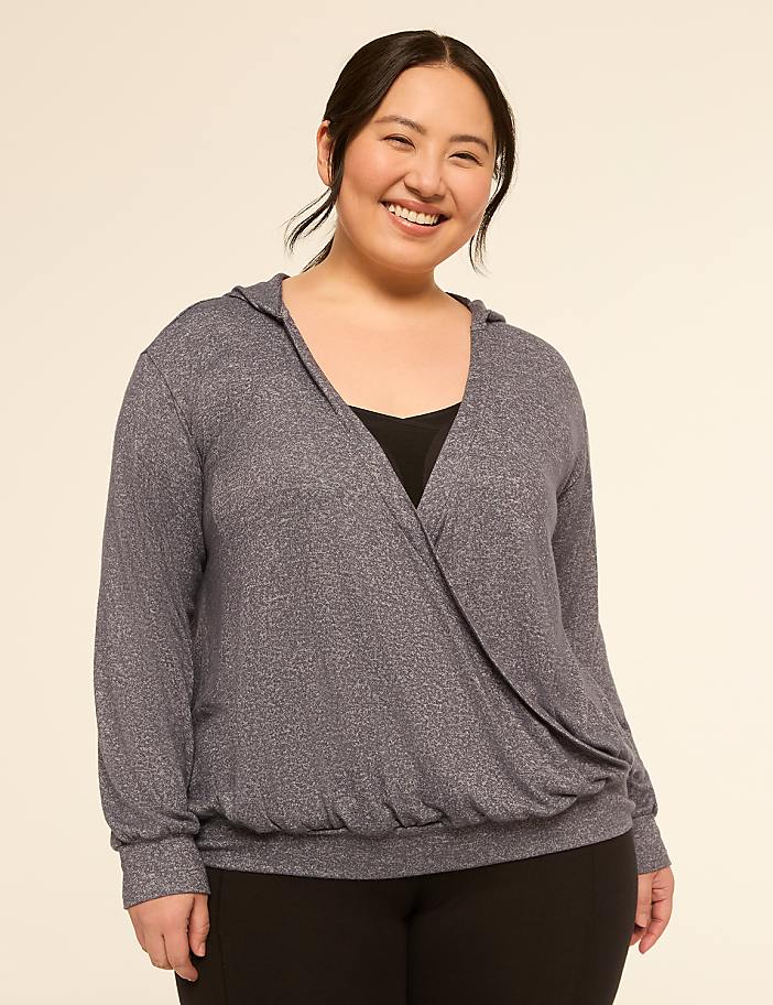 A smiling woman in a grey wrap-style hoodie stands against a plain, light-colored background.