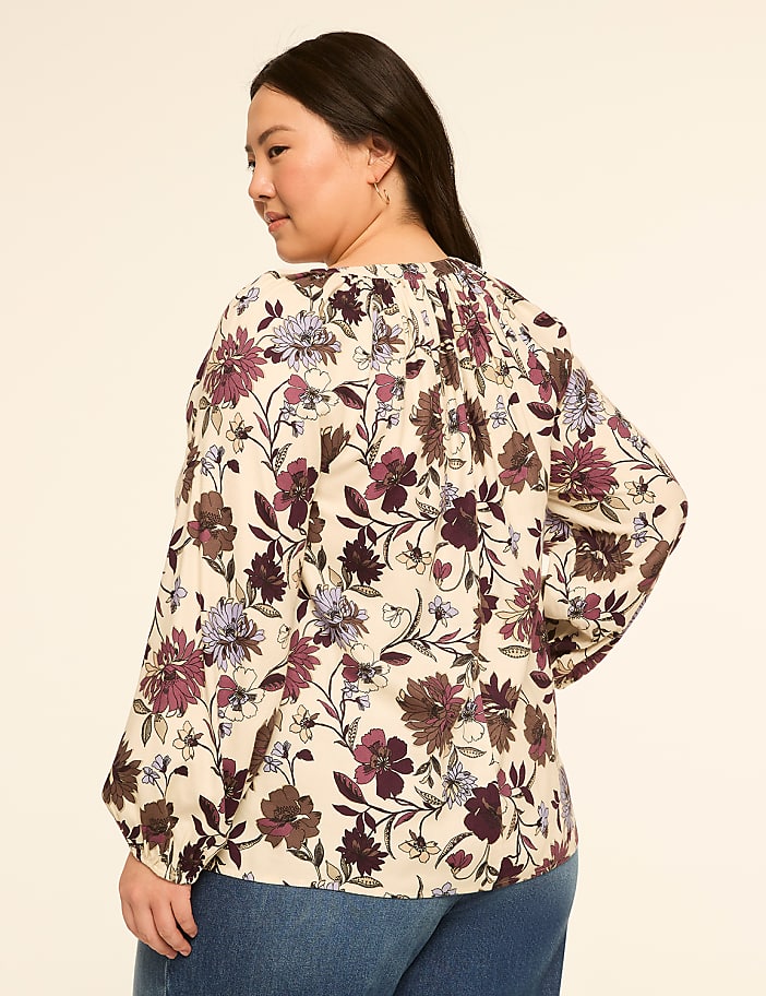 A woman in a floral blouse and jeans stands against a plain, light-colored background.