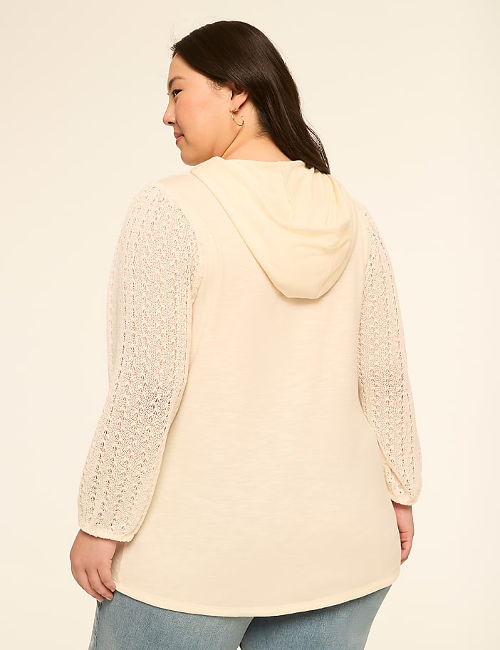 A woman in a cream-colored hooded sweater with textured sleeves stands against a plain, light-colored background.